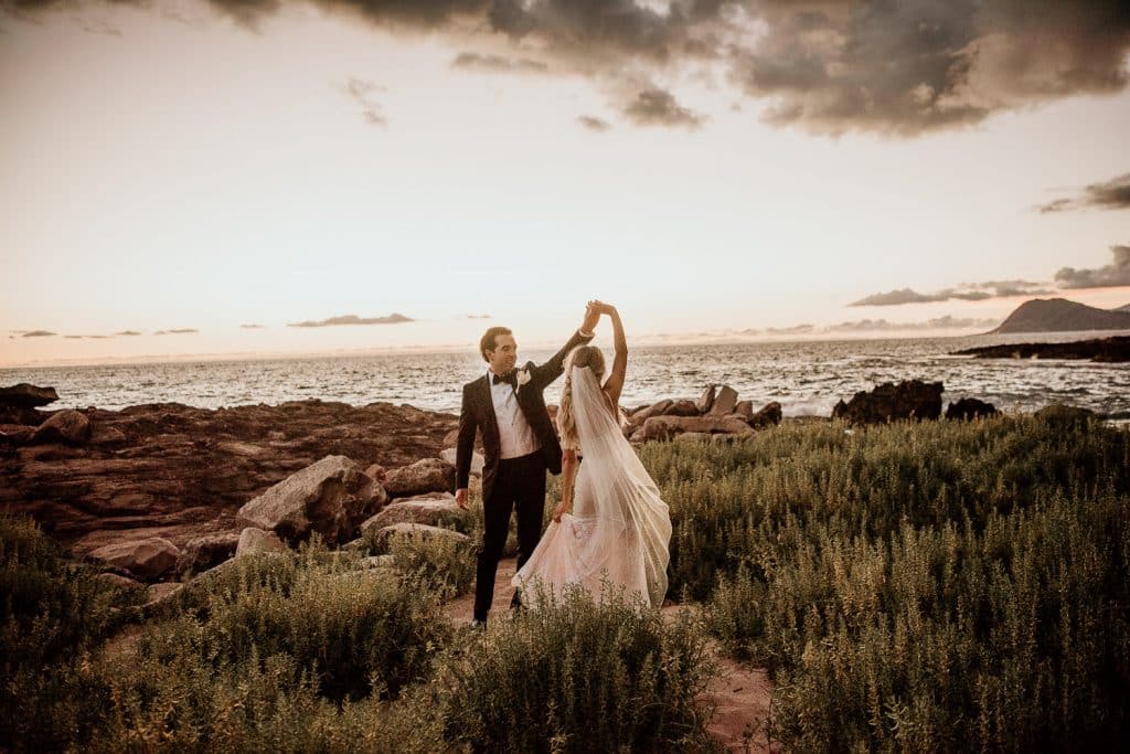 Groom in Tuxedo twirling bride around at Secret Beach in front of Four Seasons Resort Oahu at Koolina