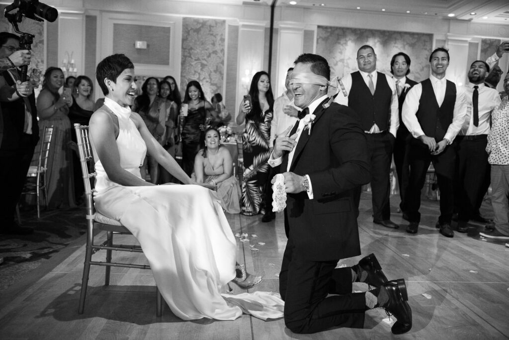 Black and White image of groom removing garter from bride while blindfolded.
