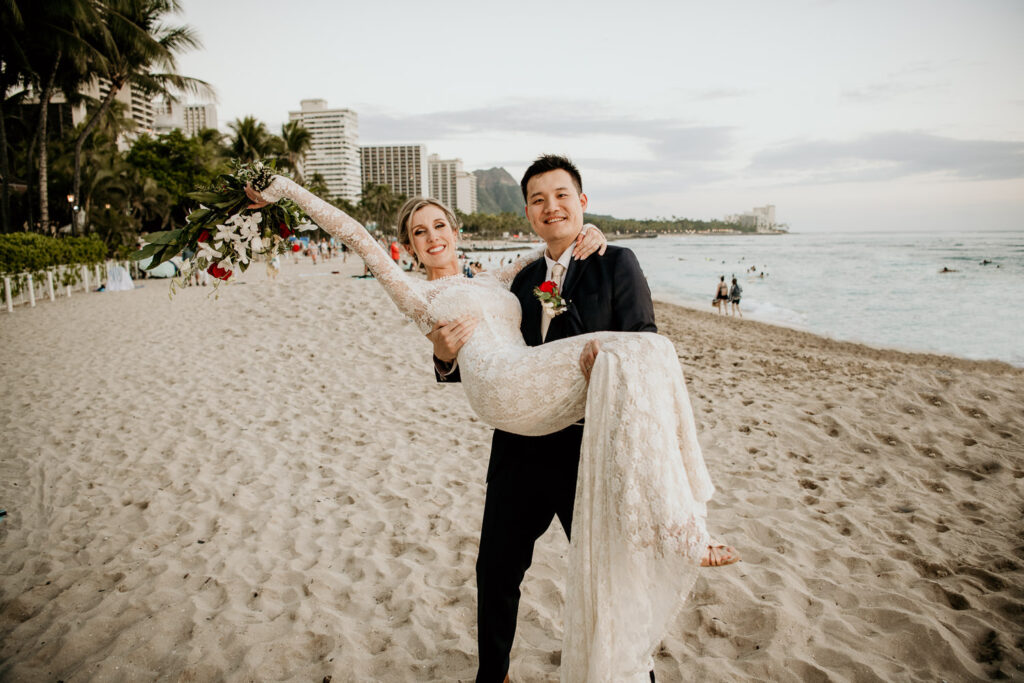 Groom carrying bride on Waikiki Beach with Diamond Head or Mt. Leahi in the background